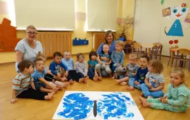 Sala zabaw-opiekunowie wraz z dziećmi siedzą na dywanie. Tuż przed nimi leży biała kartka papieru z konturem motyla wypełnionego niebieskimi odbiciami st&oacute;pek dzieci.