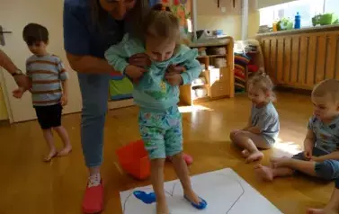 Opiekun podtrzymuje dziewczynkę, kt&oacute;ra bosymi niebieskimi st&oacute;pkami stoi na białej kartce papieru. Na niej widnieją niebieskie odbicia st&oacute;pek dzieci.