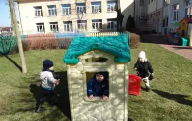 Plac zabaw-plastikowy domek a w nim dziecko wyglądające przez okienko domku. Tuż obok stoją dzieci.