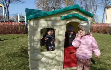 Plac zabaw-plastikowy domek a w nim dzieci. Jedno z nich wygląda przez okienko domku. Tuż obok stoją dzieci.