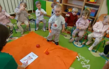 Sala zabaw-na podłodze leży pomarańczowy kocyk a na nim obrazek kr&oacute;lika, marchewka, papryka i pomidor. Tuż przy nim stoi chłopiec, w ręce trzyma marchewkę. W tle grupa dzieci siedzących na krzesełkach.