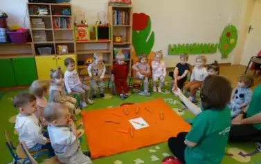 Sala zabaw-na podłodze leży pomarańczowy kocyk a na nim obrazek marchewki i warzywa marchewki. Tuż przed nim klęczy opiekun, w ręce trzyma marchewkę. W tle dzieci siedzące na krzesełkach.