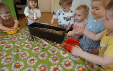 Sala zabaw-dzieci stoją przy stoliku, na kt&oacute;rym znajduje się skrzynka z ziemią i ziarenkami.
