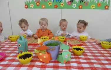 Sala zabaw-dzieci siedzą przy stoliku, na kt&oacute;rym stoją ż&oacute;łte pojemniki z ziemią, tace z ziarnem i konewka.