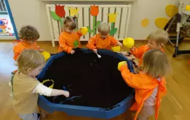 Sala zabaw-przy stoliku stoją dzieci w pomarańczowych fartuszkach. W ręce trzymają ż&oacute;łte pojemniki z ziemią.