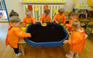 Sala zabaw-przy stoliku stoją dzieci w pomarańczowych fartuszkach. W ręce trzymają ż&oacute;łte pojemniki z ziemią.