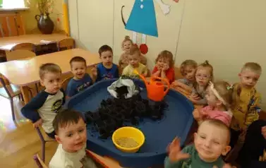 Sala zabaw-dzieci siedzą przy stoliku, na kt&oacute;rym stoją pojemniki z ziemią, miska z ziarnem i konewka. 