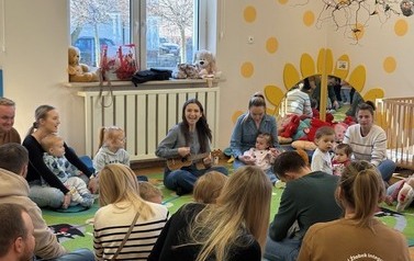 Sala zabaw-na kolorowym dywanie siedzi grupka dorosłych z dziećmi.