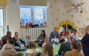 Sala zabaw-na kolorowym dywanie siedzi grupka dorosłych z dziećmi.