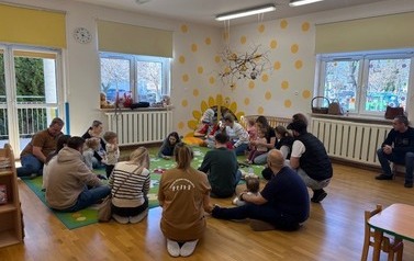 Sala zabaw-na kolorowym dywanie siedzi grupka dorosłych z dziećmi.