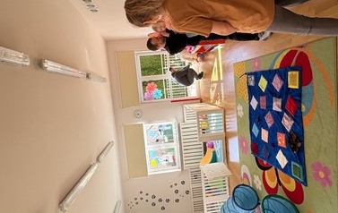 Sala zabaw-na podłodze leży kolorowa mata, w tle grupka dorosłych z dziećmi.