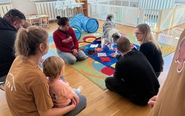 Sala zabaw-osoba dorosła siedzi z psem na podłodze a przed nimi grupka dorosłych z dziećmi siedzący na dywanie.