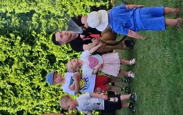 Na zielonej trawie kuca pan strażak. W ręce trzyma wąż, z kt&oacute;rego leci woda.Tuż obok grupka dzieci.