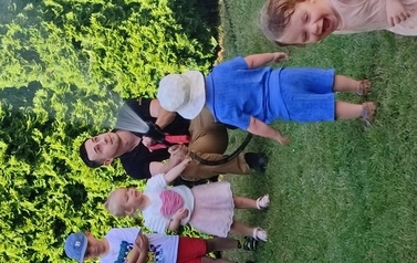 Na zielonej trawie kuca pan strażak. W ręce trzyma wąż, z kt&oacute;rego leci woda.Tuż obok grupka dzieci.