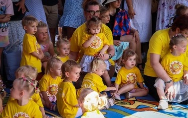 Grupka dzieci w ż&oacute;łtych koszulkach siedzi na kocu na trawie.