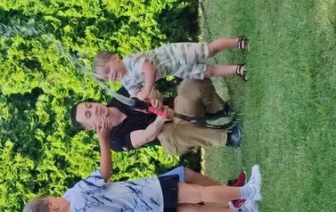 Na zielonej trawie kuca pan strażak. W ręce trzyma wąż, z kt&oacute;rego leci woda.Tuż obok grupka dzieci.