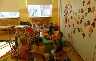 Grupa dzieci siedzi przy stoliku, na kt&oacute;rym leżą jabłka i stoi urządzenie do robienia sok&oacute;w.