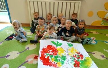 Na dywanie siedzą dzieci, tuż przed nimi leży karton z owocami.