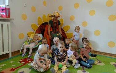 Na krzesełku siedzi Pani Jesień a wok&oacute;ł niej grupa dzieci. W tle jasna ściana a na niej ż&oacute;łte słońce.