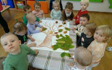 Dzieci stoją przy stoliku, na kt&oacute;rym leży karton z rysunkiem drzewa i przyklejają do niego jesienne liście.