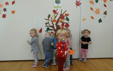 Na pierwszym planie chłopiec trzymający w ręce pomarańczowy liść, w tle jesienne drzewo i grupa dzieci.