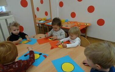 Sala zabaw - grupa dzieci siedzi przy stoliku, na kt&oacute;rym leżą kolorowe kartki papieru o r&oacute;żnych kształtach. W ręce trzymają klej, kt&oacute;rym dotykają kartek papieru.