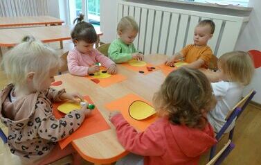 Sala zabaw - grupa dzieci siedzi przy stoliku, na kt&oacute;rym leżą kolorowe kartki papieru o r&oacute;żnych kształtach. W ręce trzymają klej, kt&oacute;rym dotykają kartek papieru.
