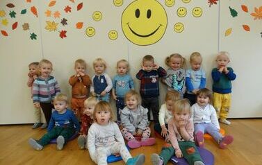 Sala zabaw - grupa dzieci stoi na tle ściany, na kt&oacute;rej wisi ż&oacute;łta buźka z szerokim uśmiechem.