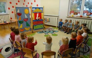 Sala zabaw - grupa dzieci siedzi na krzesełkach, przed nimi stoi kolorowy teatralny parawan, zza kt&oacute;rego wyglądają dwie kukiełki.  