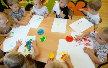 Sala zabaw - przy stoliku siedzi grupka dzieci, tuż przed nimi leżą białe kartki papieru. Na stole stoją pojemniki z kolorowymi farbami. Dzieci dotykają paluszkami farb i kartek papieru.