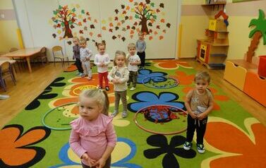Sala zabaw - grupa dzieci stoi na dywanie, na kt&oacute;rym leżą kolorowe koła a w ich wnętrzach klocki tego samego koloru. W tle jasna ściana a na niej jesienne drzewa. 