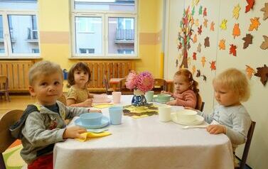 Sala zabaw - grupka dzieci siedzi przy stoliku nakrytym białym obrusem, na kt&oacute;rym stoi kolorowa zastawa stołowa.