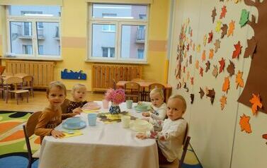 Sala zabaw - grupka dzieci siedzi przy stoliku nakrytym białym obrusem, na kt&oacute;rym stoi kolorowa zastawa stołowa.