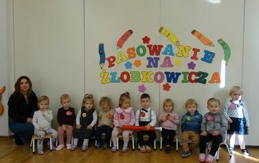 Sala zabaw - dyrektor żłobka i  grupka dzieci siedzi na krzesełkach, dwoje z nich trzyma w ręce duży oł&oacute;wek, w tle kolorowy napis "pasowanie na żłobkowicza".