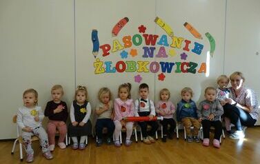 Sala zabaw - opiekun z grupą dzieci siedzi na krzesełkach, dwoje z nich trzyma w ręce czerwony duży oł&oacute;wek, w tle kolorowy napis "pasowanie na żłobkowicza".