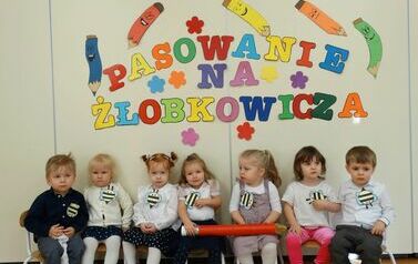 Sala zabaw - grupka dzieci siedzi na krzesełkach, dwoje z nich trzyma w ręce czerwony duży oł&oacute;wek, w tle kolorowy napis "pasowanie na żłobkowicza".