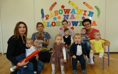 Sala zabaw - na pierwszym planie dyrektor żłobka z czerwonym oł&oacute;wkiem w ręce, w tle grupka dzieci i opiekunowie grup z dziećmi na kolanach a nad nimi kolorowy napis "pasowanie na żłobkowicza". 