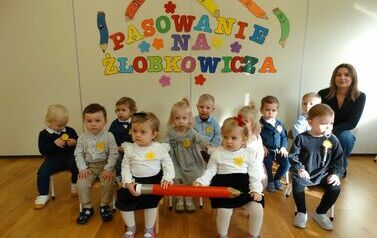 Sala zabaw - na pierwszym planie dwie dziewczynki trzymają w ręce duży czerwony oł&oacute;wek, w tle dyrektor żłobka i grupka dzieci siedzą na krzesełkach a nad nimi widnieje kolorowy napis "pasowanie na żłobkowicza". 
