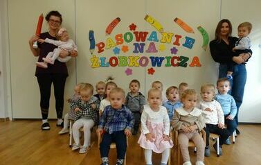 Sala zabaw - grupa dzieci siedzi na krzesełkach, w tle kolorowy napis "pasowanie na żłobkowicza". Tuż przy napisie po obydwu stronach stoją osoby dorosłe z dziećmi na ręce, jedna z nich trzyma w ręce dużą czerwoną kredkę.
