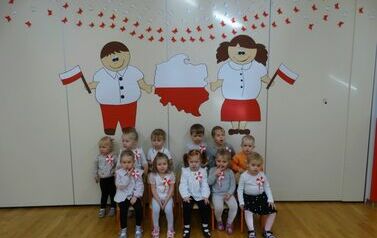 Sala zabaw - na tle jasnej ściany, udekorowanej symbolami narodowymi, siedzi grupa dzieci w odświętnych ubraniach. Ich koszulki zdobią biało-czerwone kotyliony.