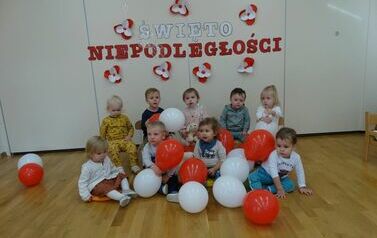 Sala zabaw - na tle jasnej ściany, na kt&oacute;rej widnieje napis Święto Niepodległości, siedzi grupa dzieci w odświętnych ubrankach. Między nimi leżą biało-czerwone balony.