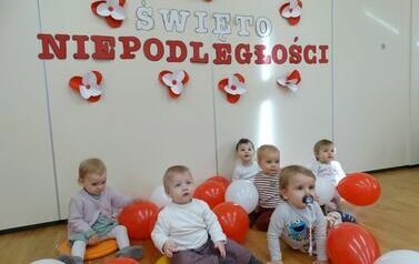 Sala zabaw - na tle jasnej ściany, na kt&oacute;rej widnieje napis Święto Niepodległości, siedzi grupa dzieci w odświętnych ubrankach. Między nimi leżą biało-czerwone balony.
