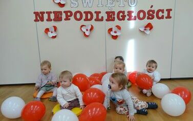 Sala zabaw - na tle jasnej ściany, na kt&oacute;rej widnieje napis Święto Niepodległości, siedzi grupa dzieci w odświętnych ubrankach. Między nimi leżą biało-czerwone balony.