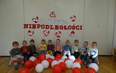 Sala zabaw - na tle jasnej ściany, na kt&oacute;rej widnieje napis Święto Niepodległości, siedzi grupa dzieci w odświętnych ubrankach. Tuż przed nimi leżą biało-czerwone balony.