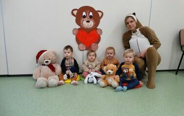 Sala zabaw - grupka dzieci z opiekunem w stroju misia siedzą na podłodze. W ręce trzymają pluszowe misie. W tle jasna ściana a na niej brązowy misio z czerwonym sercem.