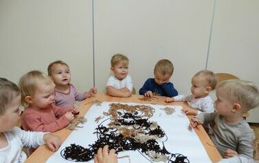 Sala zabaw - grupa dzieci siedzi przy stoliku, na kt&oacute;rym leży duży karton z rysunkiem misia. W ręce trzymają kawałki wełny, kt&oacute;rą układają na rysunku misia.