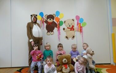 Sala zabaw - grupa dzieci siedzi na dywanie, tuż obok stoi opiekun w stroju misia. Maluszki trzymają w ręce pluszowego misia. W tle jasna ściana a na niej dwa misie z kolorowymi balonami.