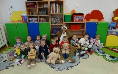 Sala zabaw - grupa dzieci z opiekunem w stroju misia siedzi na dywanie. Każde z nich trzyma w ręce pluszowego misia. W tle kolorowe szafki z zabawkami.