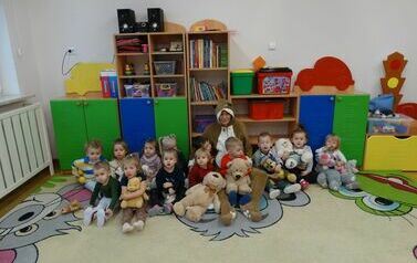Sala zabaw - grupa dzieci z opiekunem w stroju misia siedzi na dywanie. Każde z nich trzyma w ręce pluszowego misia. W tle kolorowe szafki z zabawkami.