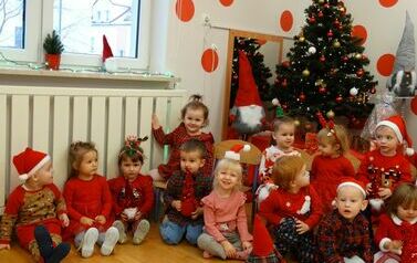 Sala zabaw - grupa dzieci w czerwonych ubrankach siedzi na podłodze, tuż za nimi błyszczy pięknie udekorowana choinka.